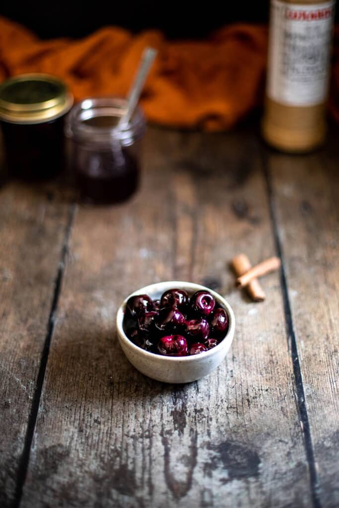 45 degree shot of small bowl of dark cherries in foreground, jar with more cherries and bottle of luxardo in the background