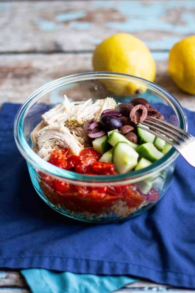 A bowl full of quinoa, shredded chicken, kalamata olives, cucumber, roasted tomatoes, and feta sauce