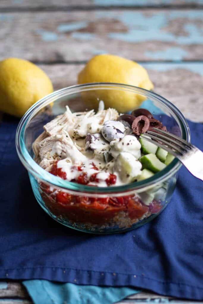 A bowl full of quinoa, shredded chicken, kalamata olives, cucumber, roasted tomatoes, and feta sauce