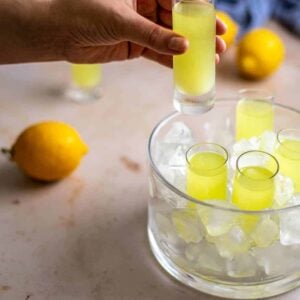 a glass dish with tall sides is filled with ice and has 4 shot glasses filled with limoncello; a hand is reaching in from the left and holds another shot glass with limoncello