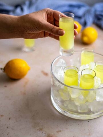 a glass dish with tall sides is filled with ice and has 4 shot glasses filled with limoncello; a hand is reaching in from the left and holds another shot glass with limoncello