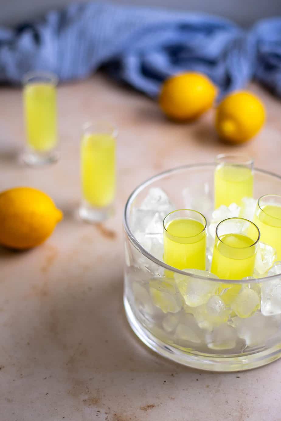 a glass dish is filled with ice and holds 4 tall shot glasses of limoncello; two shot glasses are in the background, along with lemons