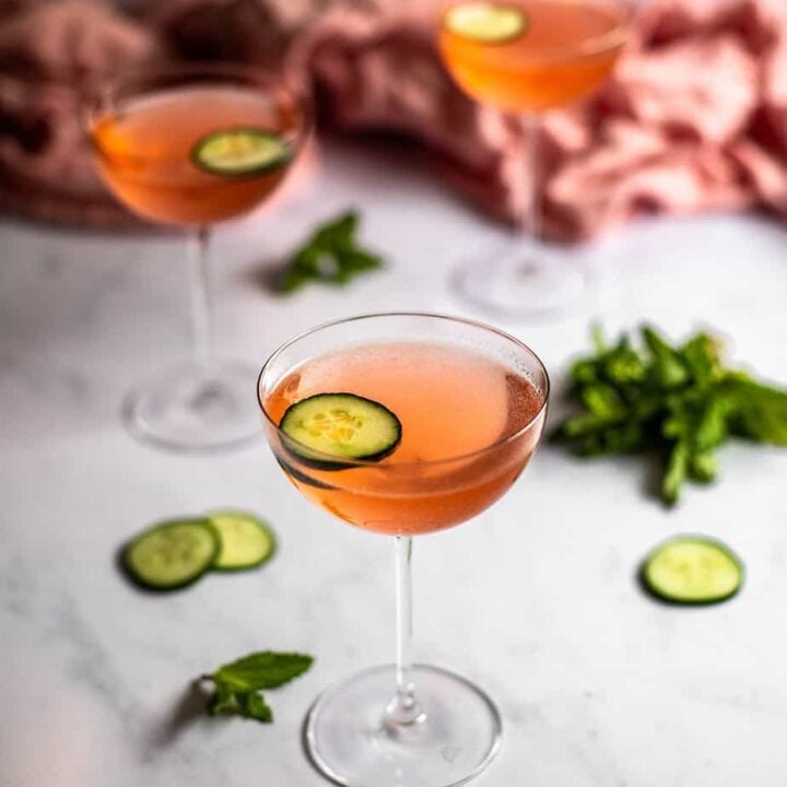 three coupe glasses on a white marble slab, glasses are filled with pink liquid and are garnished with a cucumber slice