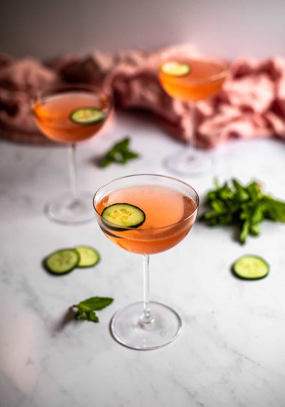 three coupe glasses on a white marble slab, glasses are filled with pink liquid and are garnished with a cucumber slice