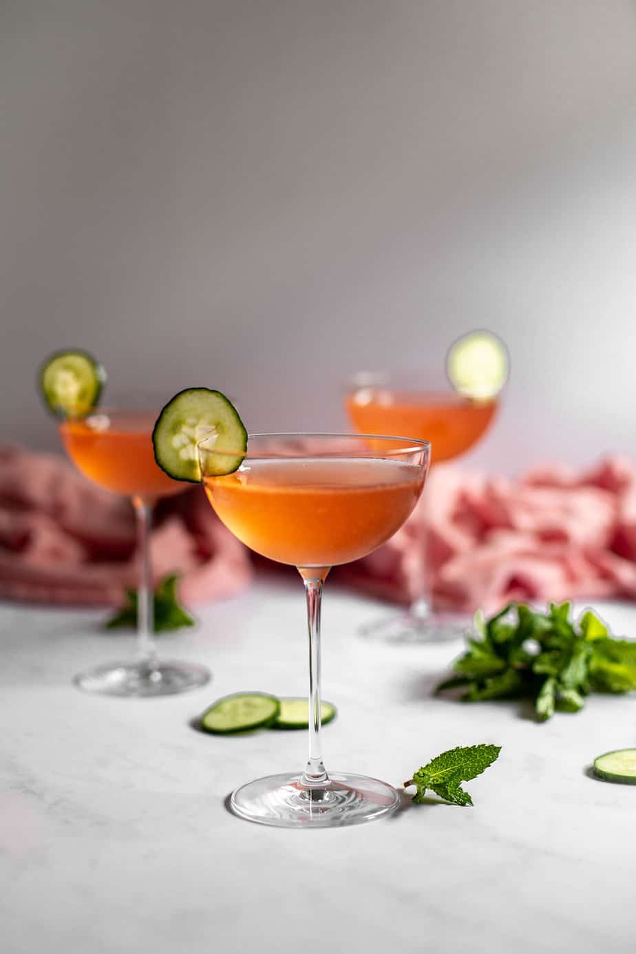 a straight on view of three coupe glasses filled with pink liquid and garnished with cucumber slices