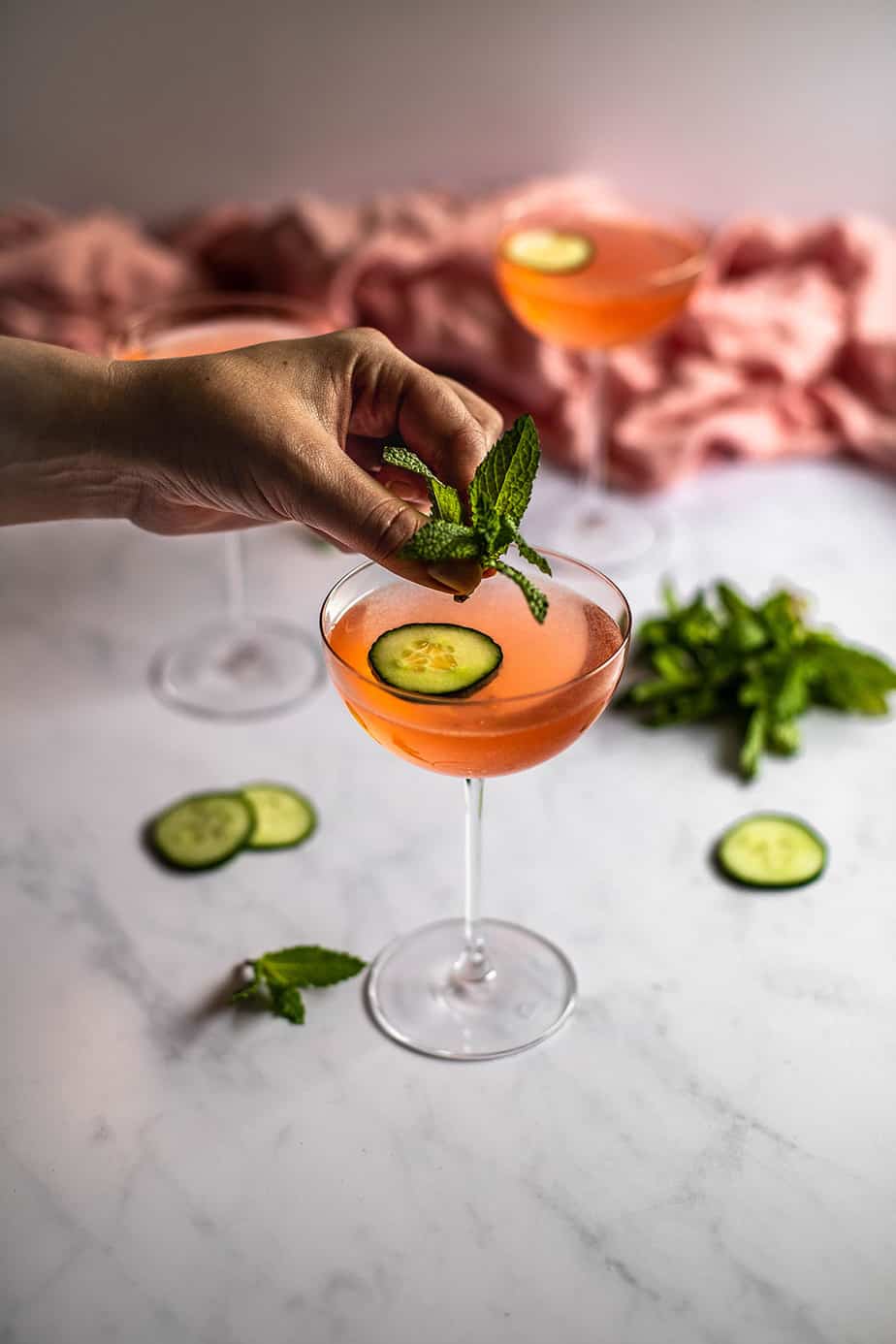 a hand getting ready to place a sprig of fresh mint into a couple glass filled with pink liquid and garnished with a cucumber slice