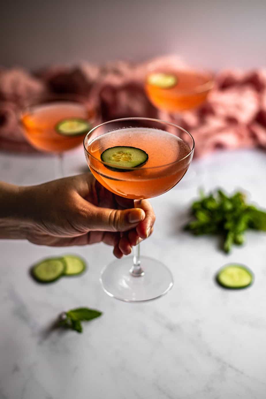 a hand holding a coupe glass filled with pink liquid and garnished with a cucumber slice