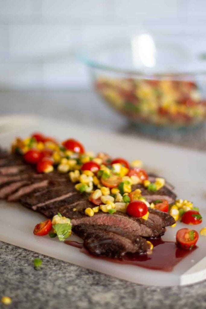 flank steak, sliced, laying on a white cutting board, topped with corn salsa