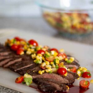 flank steak, sliced, laying on a white cutting board, topped with corn salsa