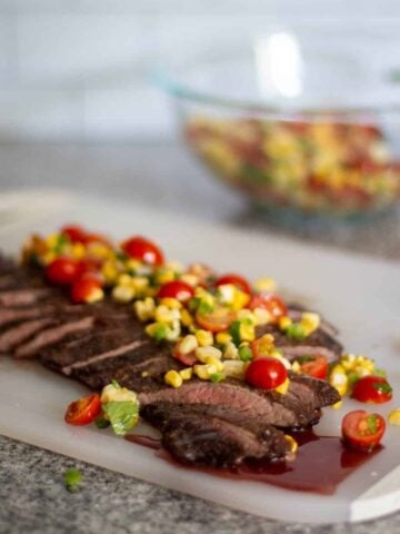 flank steak, sliced, laying on a white cutting board, topped with corn salsa