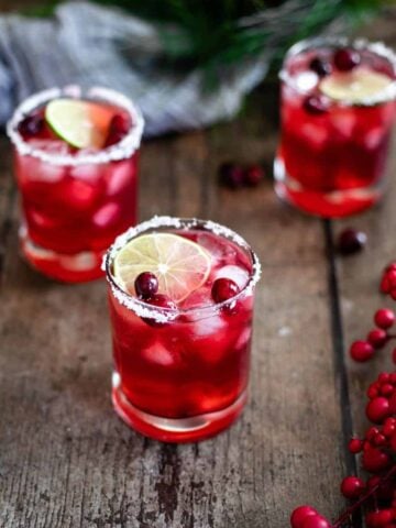 three short glasses filled with red cranberry margaritas; glasses are garnished with a salt rim, a slice of lime, and fresh cranberries; the glasses are sitting on a warm wood backdrop