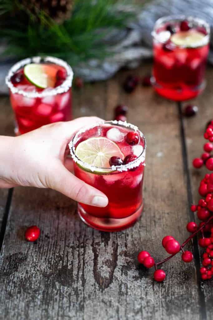three rocks glasses, filled with bright red cranberry margaritas; garnished with a slice of lime and several fresh cranberries; a hand entered the frame from the left to grab one of the glasses