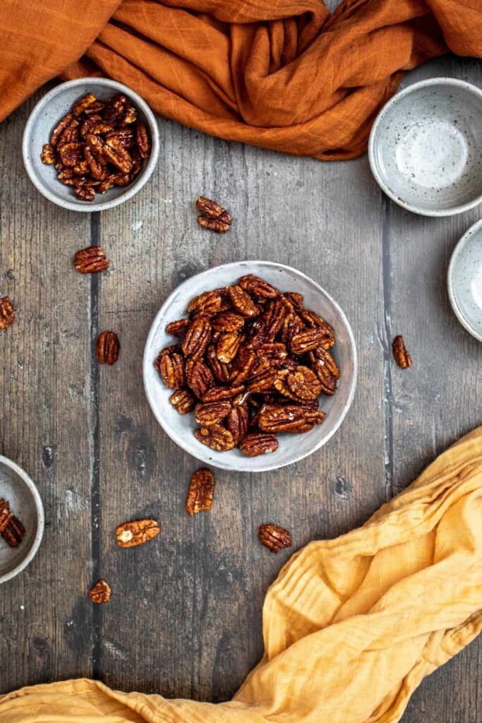 a medium-sized bowl of candied pecans in the center, 1 smaller bowl of nuts above and to the left, and 1 small empty bowl, on a dark wooden background