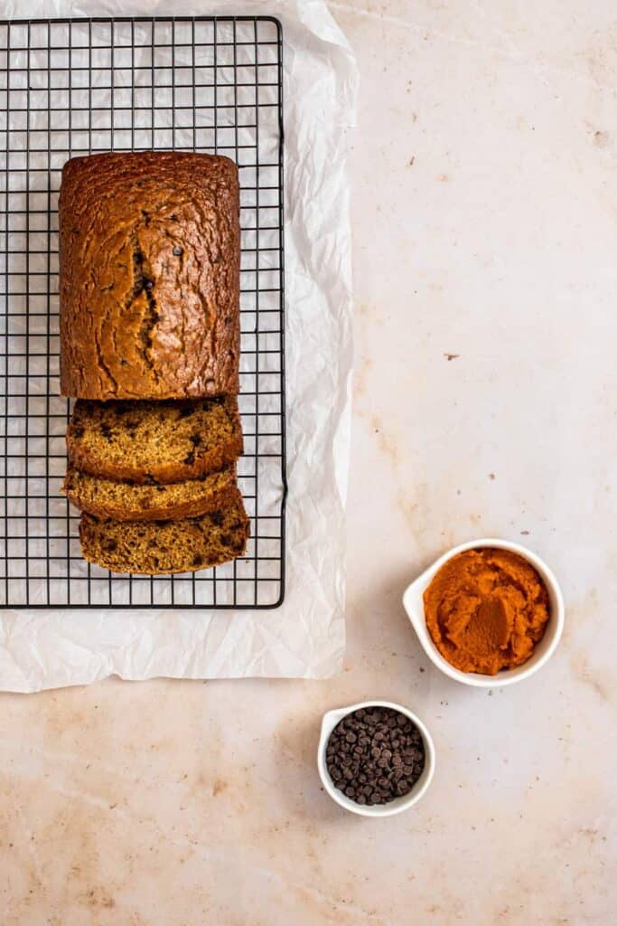 photographed from above, a loaf of pumpkin banana bread with three slices, sitting on a wire baking rack, with small bowls of pumpkin puree and chocolate chips set to the bottom right of the photo