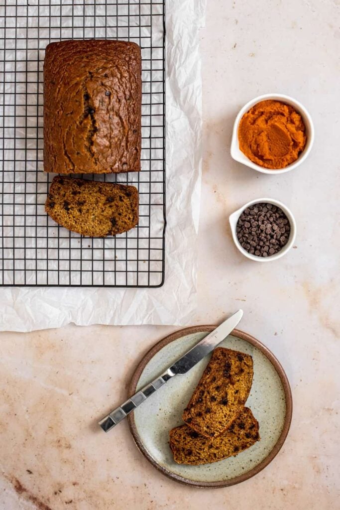 viewed from above, a loaf of pumpkin banana bread on a wire baking rack with one cut slice on the rack, two slices on a plate with a knife, and pumpkin puree and chocolate chips in small bowls set off to the side