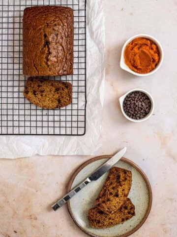 viewed from above, a loaf of pumpkin banana bread on a wire baking rack with one cut slice on the rack, two slices on a plate with a knife, and pumpkin puree and chocolate chips in small bowls set off to the side