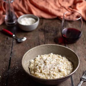 bowl of cacio e pepe risotto with glass of wine, dish of cheese, and pepper mill in background