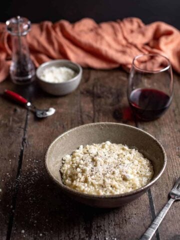 bowl of cacio e pepe risotto with glass of wine, dish of cheese, and pepper mill in background