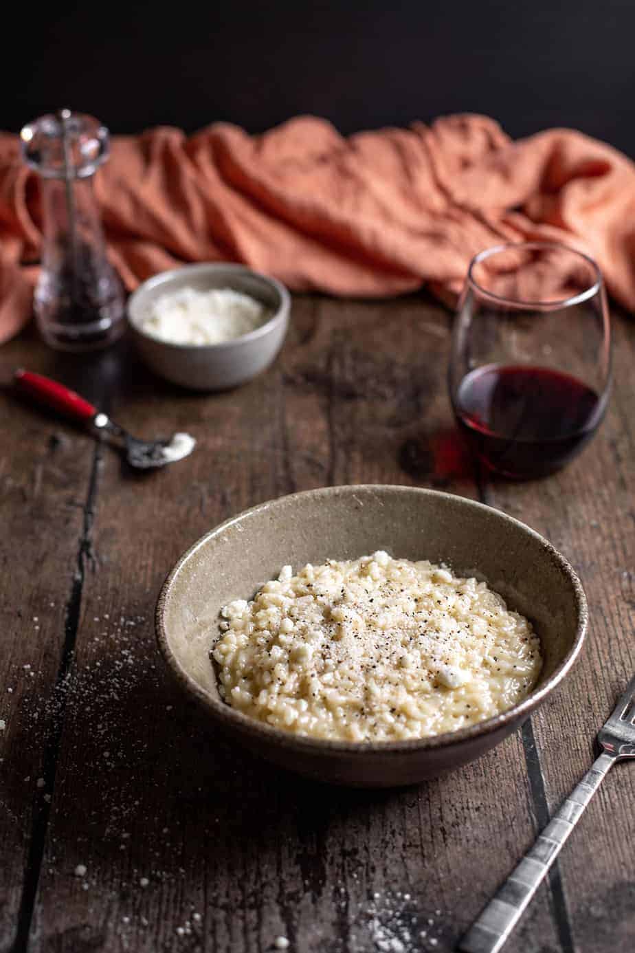 bowl of cacio e pepe risotto with glass of wine, dish of cheese, and pepper mill in background