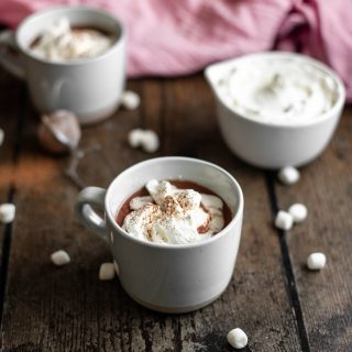 a mug of red wine hot chocolate with whipped cream and cocoa powder in the foreground, another mug of hot chocolate and a bowl of whipped cream in the background