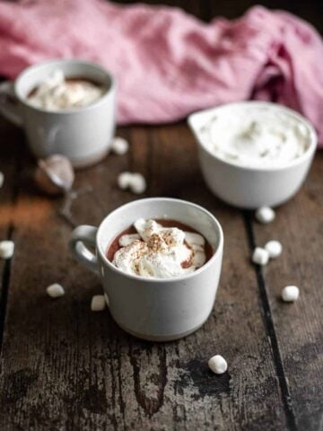 a mug of red wine hot chocolate with whipped cream and cocoa powder in the foreground, another mug of hot chocolate and a bowl of whipped cream in the background