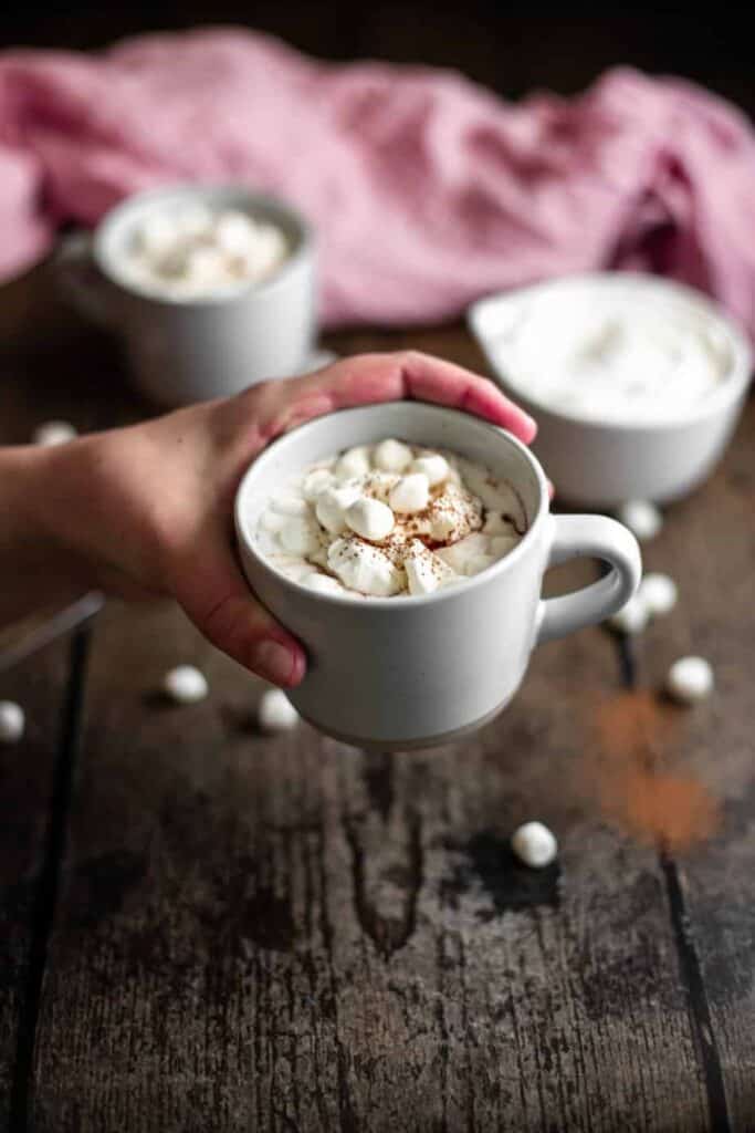 a hand holding a mug of red wine hot chocolate in the foreground, another mug and a bowl full of whipped cream in the background