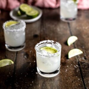 three margaritas on a dark wooden background, a dish of lime wedges in the left rear