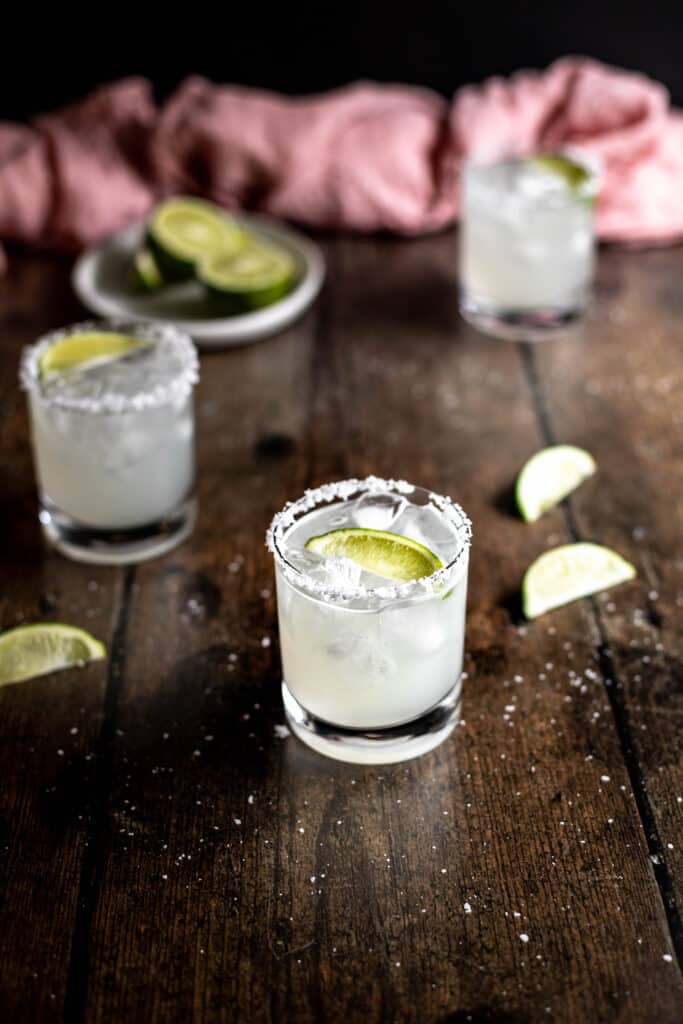 three margaritas on a dark wooden background, a dish of lime wedges in the left rear