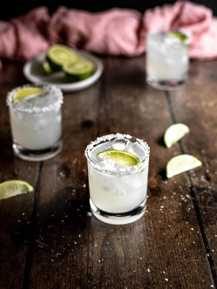 three margaritas on a dark wooden background, a dish of lime wedges in the left rear