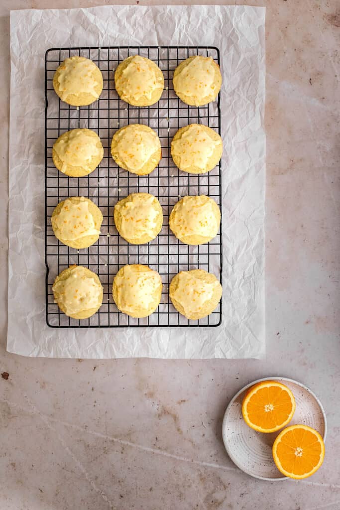 an overhead view of 12 orange cookies on a wire cooling rack, a dish with halved oranges in the lower righthand corner