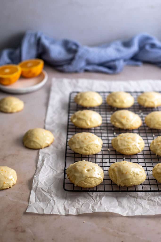 45 degree angle view of orange cookies on a wire cooling rack, 3 cookies to the left not on the rack, halved oranges in background