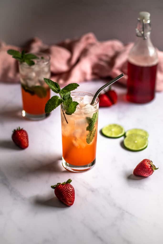 a 45 degree angle photo of two strawberry mojitos, a bottle with red strawberry simple syrup in the background