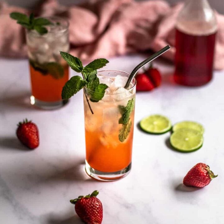 a 45 degree angle photo of two strawberry mojitos, a bottle with red strawberry simple syrup in the background