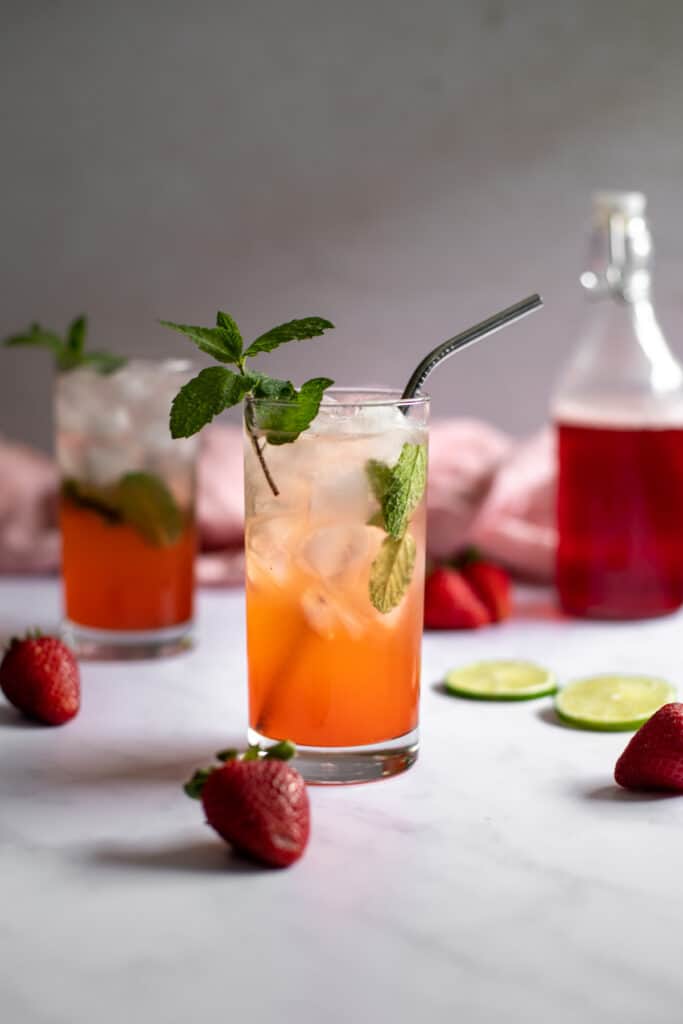 a head on photo of two strawberry mojitos, a bottle with red strawberry simple syrup in the background