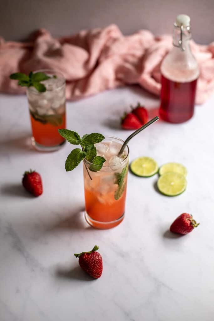 a 45 degree angle photo of two strawberry mojitos, a bottle with red strawberry simple syrup in the background