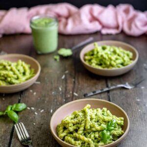 three small bowls containing short cut pasta with pesto sauce, small jar of broccoli pesto in background