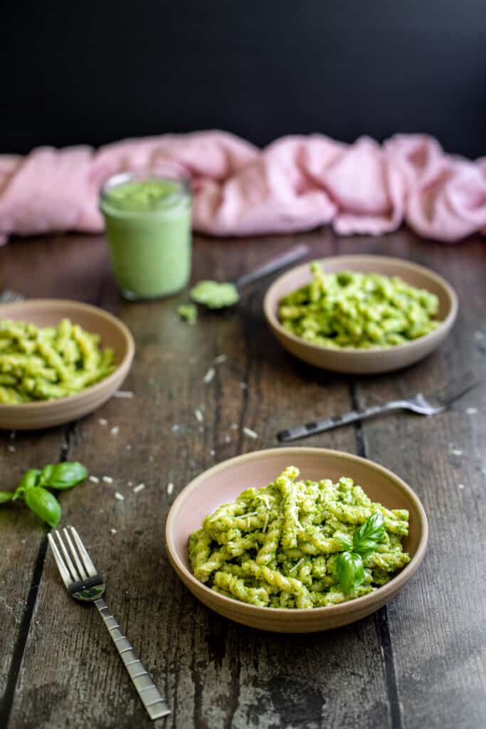 three small bowls containing short cut pasta with pesto sauce, small jar of broccoli pesto in background