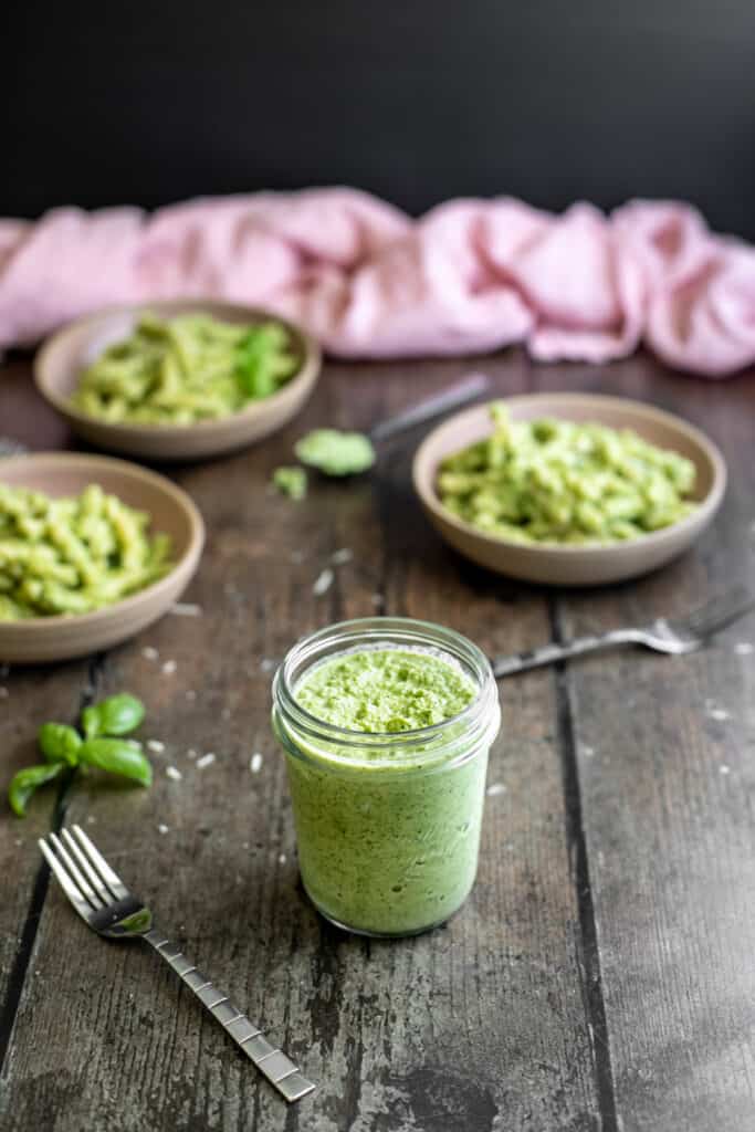 small jar of broccoli pesto, three bowls of short cut pasta with pesto in background