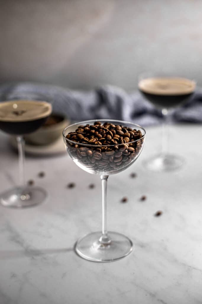 a coupe glass filled with coffee beans in the foreground, two coupe glasses filled with espresso martinis in the background