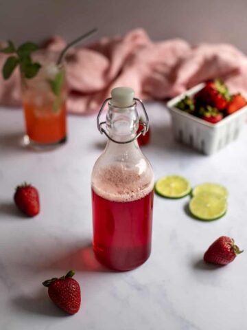 swing-top bottle full of red liquid in foreground, cocktail and small basket of strawberries in background