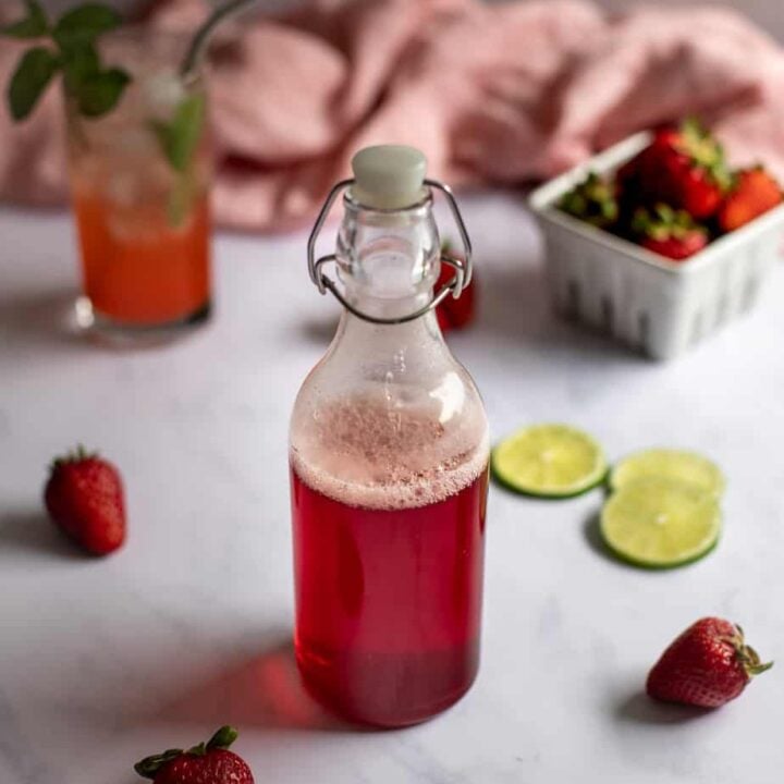swing-top bottle full of red liquid in foreground, cocktail and small basket of strawberries in background