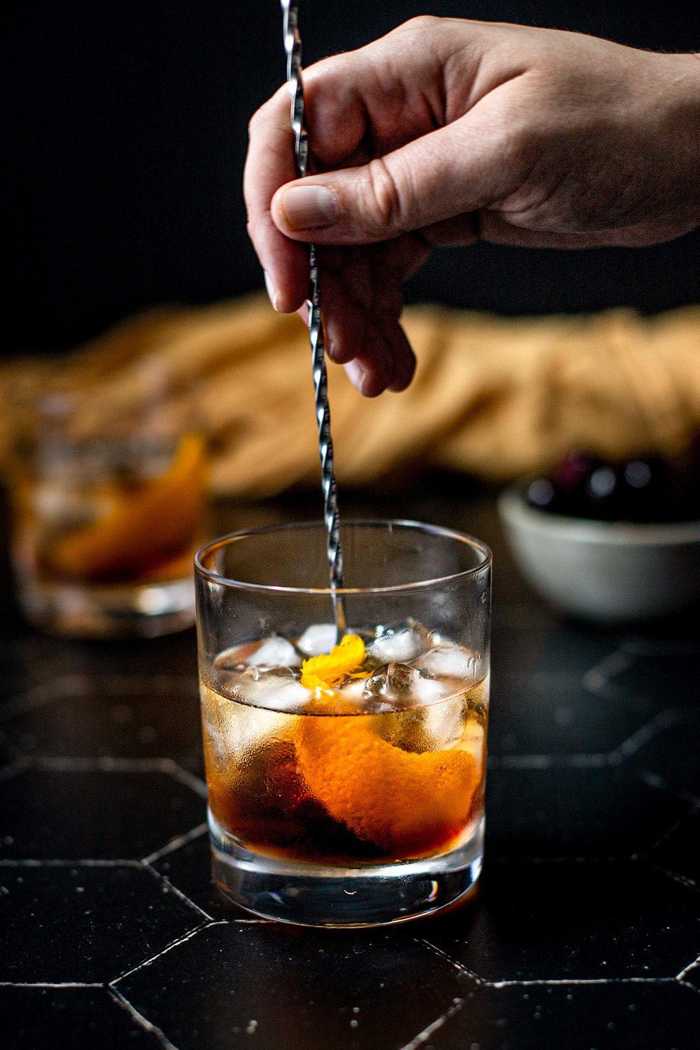 a maple old fashioned on a black tile background, a hand is stirring the cocktail with a bar spoon