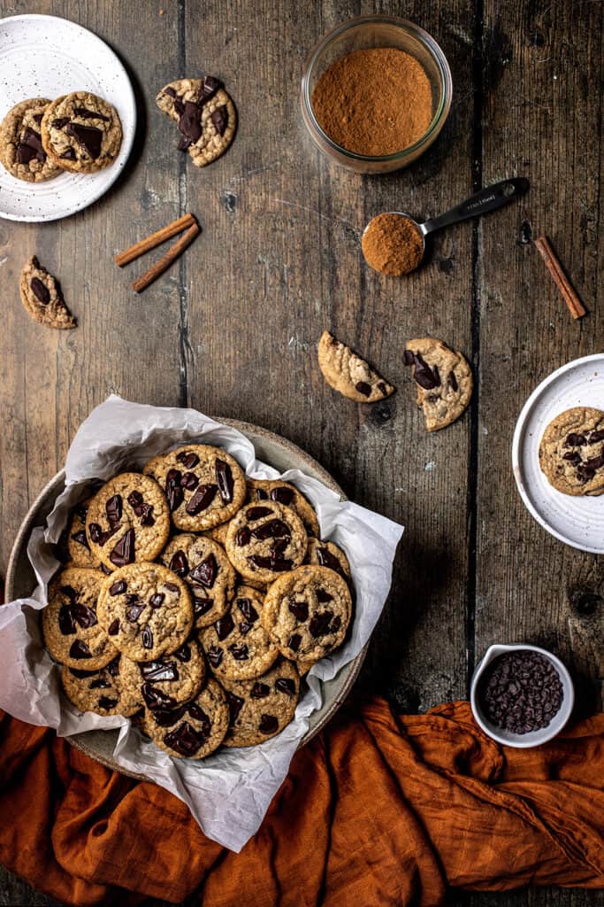 an overhead view of a bowl of pumpkin spice chocolate chip cookies, a dish of pumpkin spice, and plates of cookies