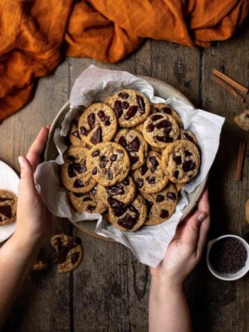 an overhead view of two hands holding a bowl of pumpkin spice chocolate chip cookies