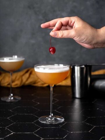 a tequila sour in a coupe glass, a hand holding a cherry above the glass
