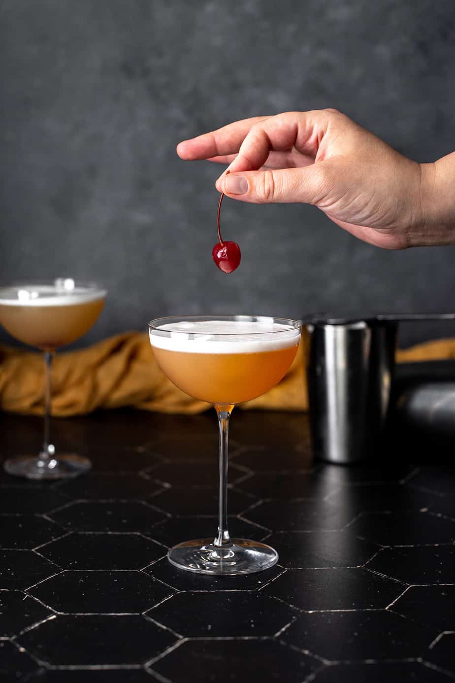 a tequila sour in a coupe glass, a hand holding a cherry above the glass