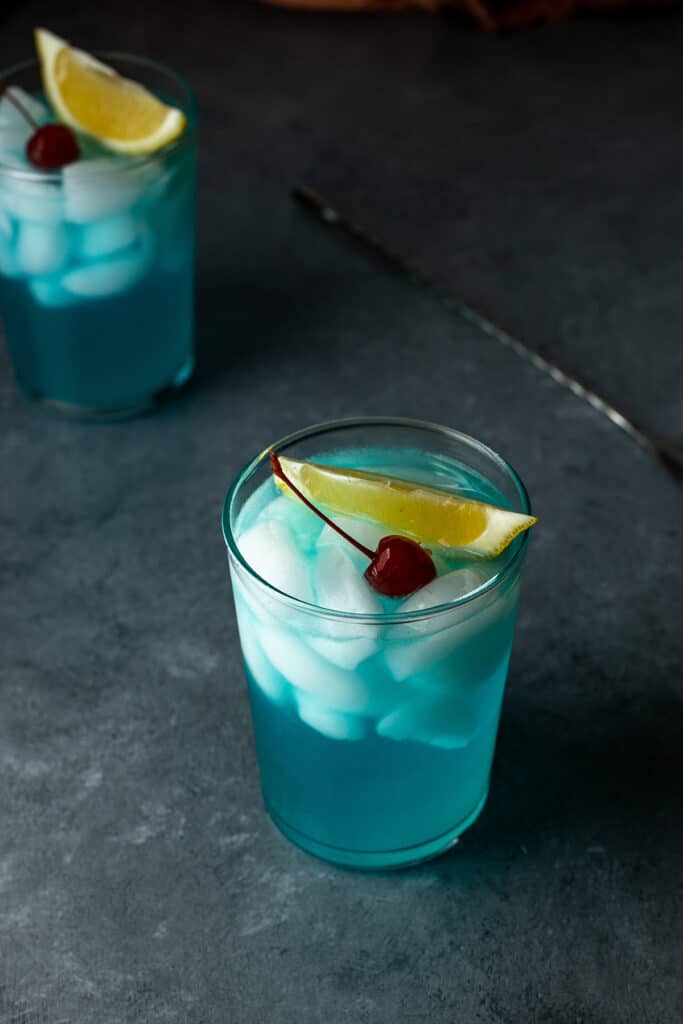 a 45 degree angle view of two glasses filled with light blue liquid, ice, and garnished with lemon and a cherry