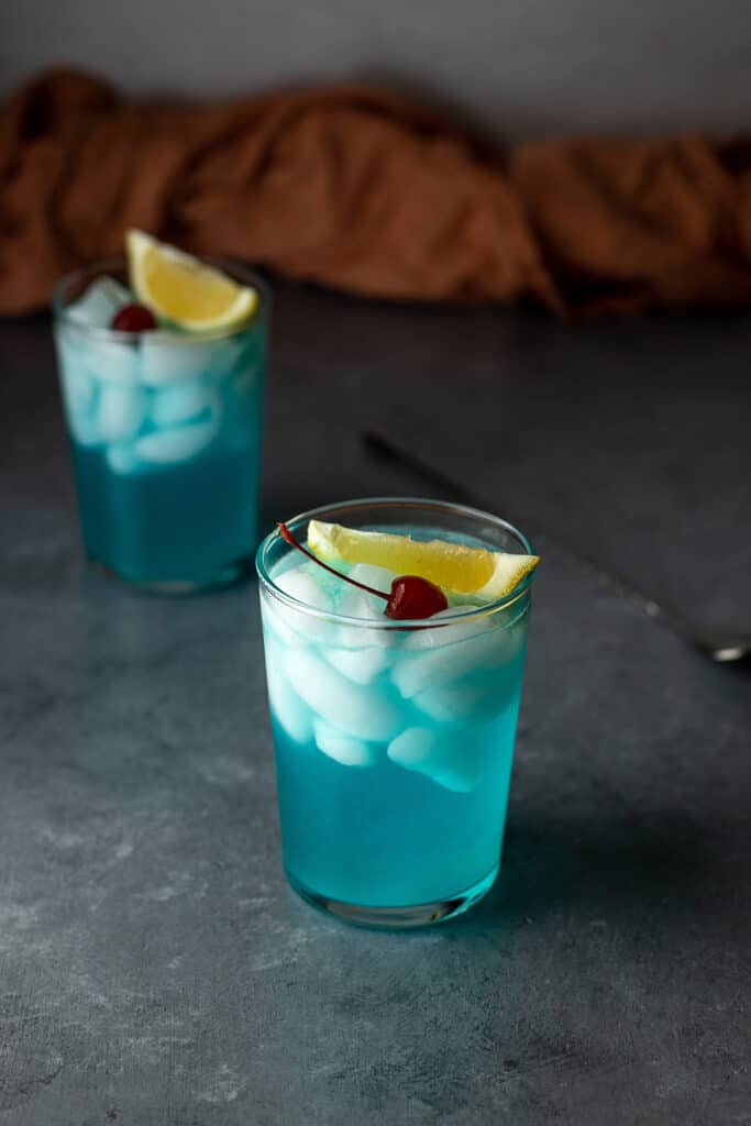 two glasses filled with light blue liquid, ice, and garnished with lemon and a cherry