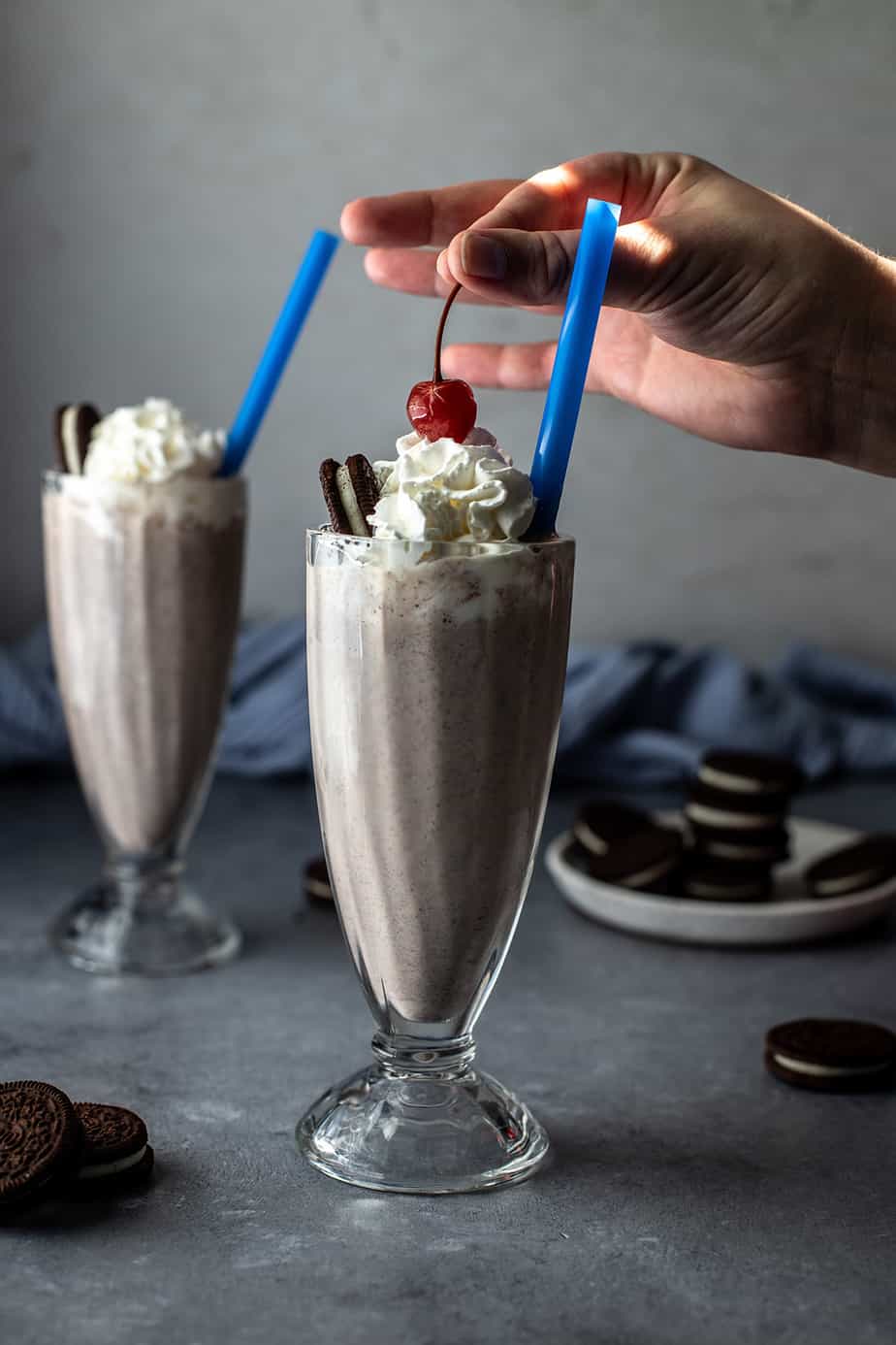 two glasses with cookies and cream milkshakes, a hand is placing a cherry on the top of one