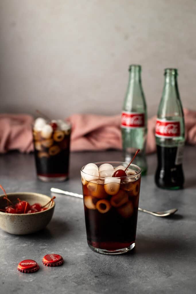 a glass full of a roy rogers drink in the foreground, another in the background along with two glass bottles of coke